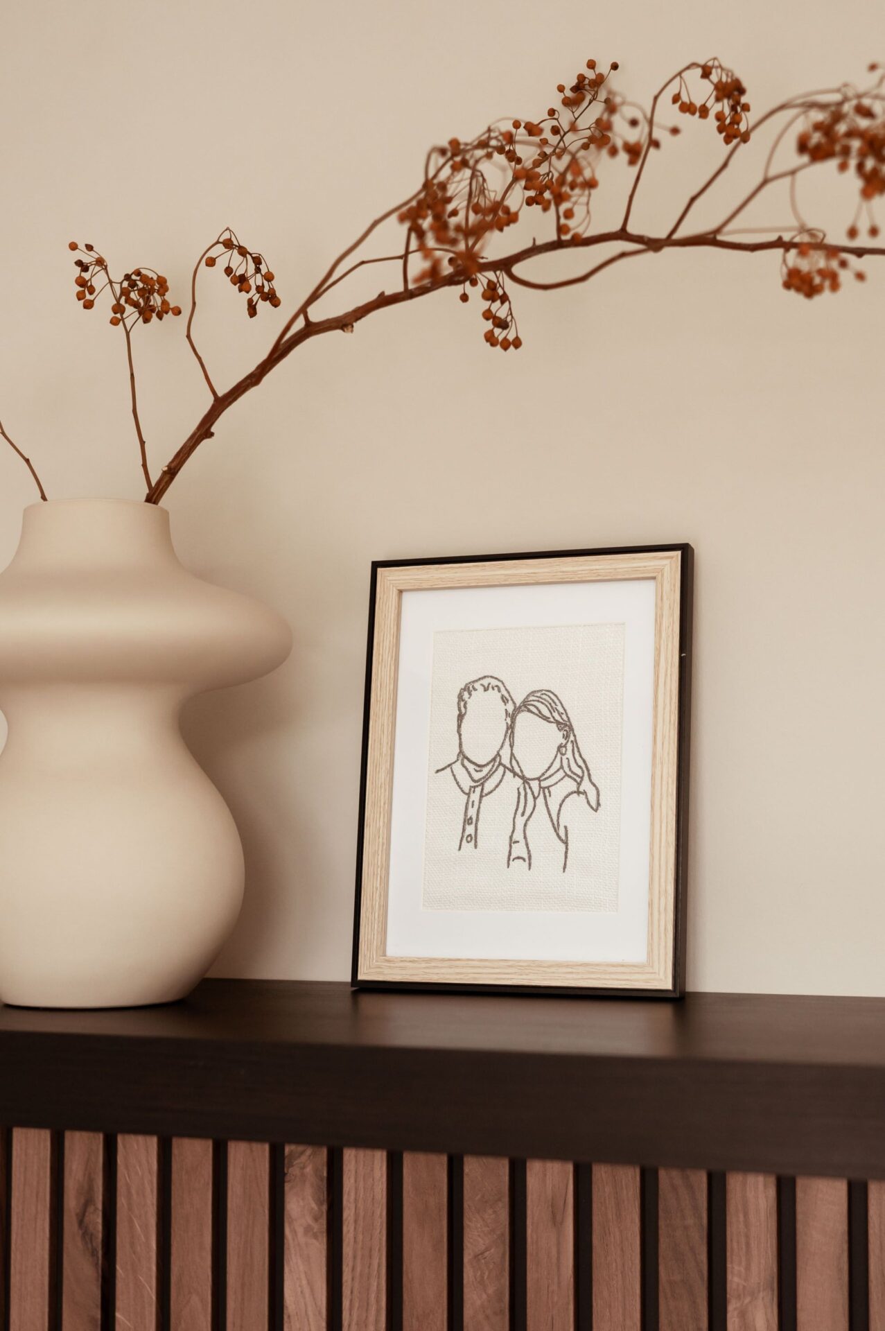 Minimalistisch interieur met gedroogde takken in crèmekleurige vaas en ingelijste lijntekening van koppel op plank